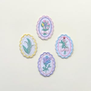 May include: Four embroidered oval patches with floral designs. Each patch features a different flower arrangement in pastel colours, including pink, blue, yellow, and green. The patches have scalloped edges in coordinating colours, creating a delicate and vintage aesthetic.
