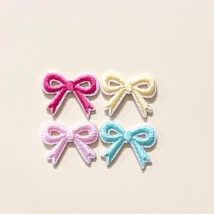 May include: Four embroidered bow patches arranged in a square grid. The top left bow is dark pink, the top right is pale yellow, the bottom left is light pink, and the bottom right is light blue. Each bow features a white stitched border.