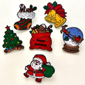 May include: A selection of festive Christmas enamel pins. The designs feature a stocking, bells, a Christmas tree, a bag with "Happy Holidays" text, a snow globe with Santa, and Santa Claus with a sack.