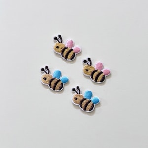May include: Four embroidered bee patches featuring gold and brown striped bodies with black antennae. Two patches have light pink wings, and two patches have light blue wings. Each bee is outlined in white thread against a plain background.