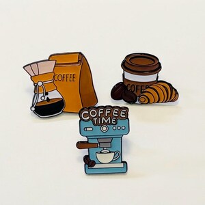 Op de afbeelding: Drie emaille pins met koffiethema's. Eén pin toont een glazen koffiezetapparaat naast een gele zak met het opschrift COFFEE. Een andere toont een bruine koffiekop met een croissant en bonen. De derde is een blauwe espressomachine met de tekst COFFEE TIME.