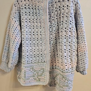 Può includere: Un cardigan di colore azzurro chiaro lavorato a uncinetto con un design sfumato rosa e verde. Il cardigan ha una chiusura con bottoni e un bordo lavorato a uncinetto.