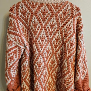 May include: A crocheted sweater with a geometric pattern in shades of orange and white. The sweater has long sleeves and a relaxed fit.