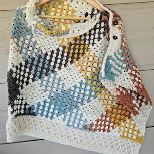 Op de afbeelding: Een gebreid poncho met een rolkraag en een geometrisch patroon in tinten bruin, blauw, wit en geel. De poncho heeft een knoopsluiting op de schouder.