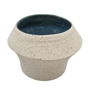 May include: A small, off-white ceramic vase with a textured surface and a dark blue interior. The vase has a unique, rounded shape with a wide opening. The exterior has a speckled appearance.
