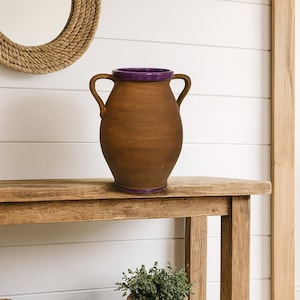 May include: A brown ceramic vase with two handles and a purple rim sits on a wooden table. A woven rope mirror hangs on the white shiplap wall behind the vase. Decorative baskets and a potted plant are on the shelf below.