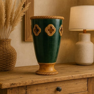May include: A dark green decorative vase with a light brown base and rim, featuring a rope-like texture. The vase has four clover-shaped cutouts. It sits on a wooden table, next to a lamp and a woven basket.