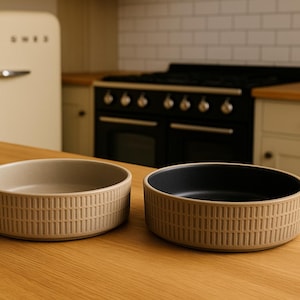 May include: Two beige ceramic bowls with a textured, vertical line pattern. One bowl has a beige interior, the other a black interior. The bowls are on a wooden surface, with a retro-style fridge and cooker in the background.