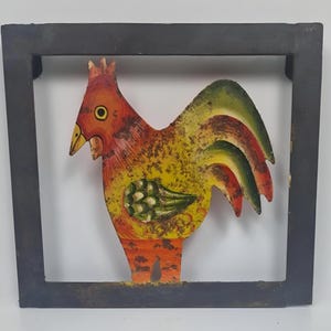 May include: A framed wooden rooster sculpture. The rooster is painted in vibrant colours: red, orange, yellow, and green, with a distressed finish. The frame is dark grey. Likely for wall decor.