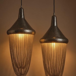 May include: Two pendant lights with a hammered metal shade and a cascade of chain fringe. The lights hang from black cords and emit a warm glow. The fixtures have a unique, elegant design.