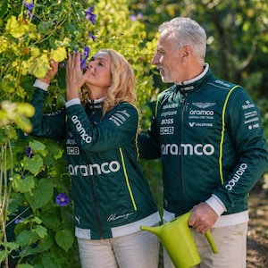 May include: A couple in dark green jackets with yellow detailing and Aramco branding. The jackets feature sponsor logos. The woman is smelling purple flowers, and the man holds a green watering can. The jackets are part of a Formula One team's apparel.