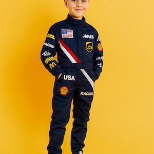 K&ouml;nnte beinhalten: Ein marineblauer Rennanzug mit wei&szlig;en und roten Akzenten, mit der amerikanischen Flagge, dem Namen "JAMES" und Logos. Der Anzug enth&auml;lt das Wort "USA" und "RACING". Das Kind tr&auml;gt wei&szlig;e Turnschuhe.