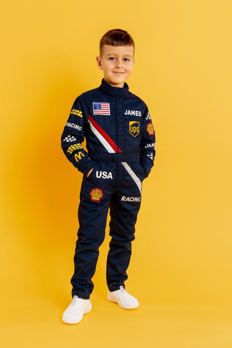K&ouml;nnte beinhalten: Ein marineblauer Rennanzug mit wei&szlig;en und roten Akzenten, mit der amerikanischen Flagge, dem Namen "JAMES" und Logos. Der Anzug enth&auml;lt das Wort "USA" und "RACING". Das Kind tr&auml;gt wei&szlig;e Turnschuhe.