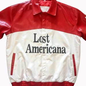 May include: A red and white jacket with the text "Lost Americana" in black. The jacket has a collared neckline, button closure, and two red pocket accents. The top portion is red, and the bottom is white.
