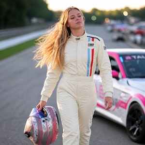 May include: A cream-coloured racing suit with blue and red stripes. The suit has the OMP logo on the chest and shoulders. A racing helmet with pink and silver accents is held in the hand. A pink and white race car is in the background.