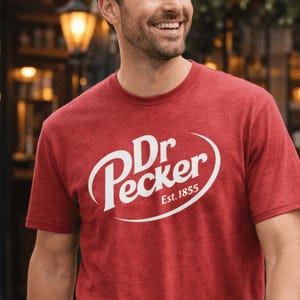 May include: Red t-shirt with the text "Dr. Pecker Est. 1855" in a white, stylized font. The shirt is short-sleeved and has a crew neck. The person wearing the shirt is smiling.