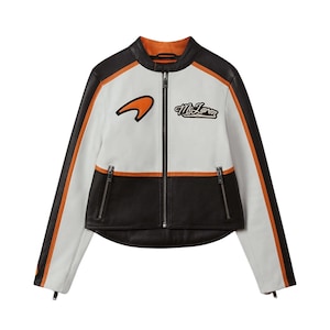 Può includere: Giacca in pelle bianca, nera e arancione con chiusura a zip. La giacca presenta il logo e la scritta McLaren sul petto, con strisce arancioni sulle maniche. La giacca ha due tasche con cerniera.