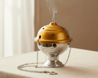 May include: A metallic incense burner featuring a silver-toned pedestal base and a removable gold-toned dome lid with circular ventilation holes. A thin silver chain is attached to the sides of the lid, with wisps of smoke rising from the top.