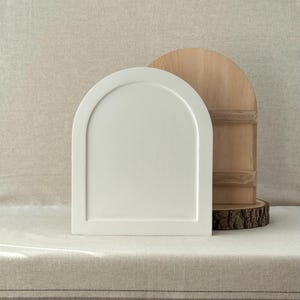 May include: A white, arched decorative tray stands in front of a wooden, half-circle cutting board. The tray has a raised border and a smooth surface. The cutting board is made of light-coloured wood and rests on a wood slice.