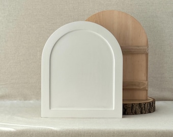 May include: A white, arched decorative tray stands in front of a wooden, half-circle cutting board. The tray has a raised border and a smooth surface. The cutting board is made of light-colored wood and rests on a wood slice.