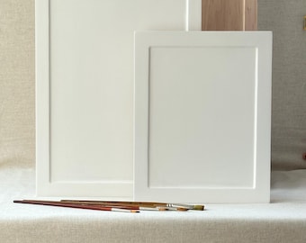 May include: Two white framed panels of different sizes stand in front of a light wood panel. Several paint brushes are arranged in front of the panels. The panels and brushes are on a neutral-toned surface.