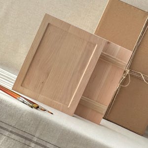 May include: Two unfinished wooden cabinet doors with a rectangular recessed panel design. A paintbrush with orange, yellow, and brown bristles rests on a striped fabric. A cardboard box tied with twine is in the background.