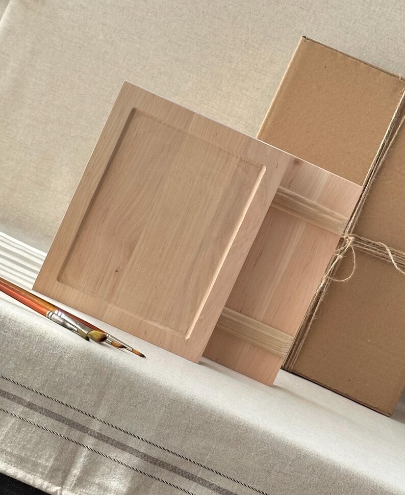 May include: Two unfinished wooden cabinet doors with a rectangular recessed panel design. A paintbrush with orange, yellow, and brown bristles rests on a striped fabric. A cardboard box tied with twine is in the background.