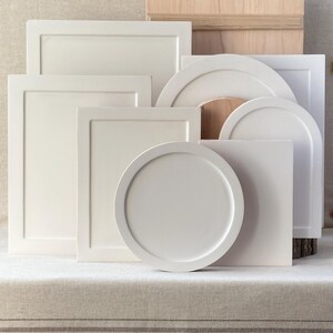 May include: A collection of off-white, geometric wooden shapes, including rectangles, squares, and circles. Some pieces have raised edges, and a wooden box is visible in the background. The items are arranged on a neutral-toned surface.