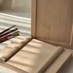 May include: Two unfinished wooden panels in a light brown hue. One panel features a recessed square, and the other has decorative trim. Paintbrushes are visible in the background, suggesting an art or craft theme.