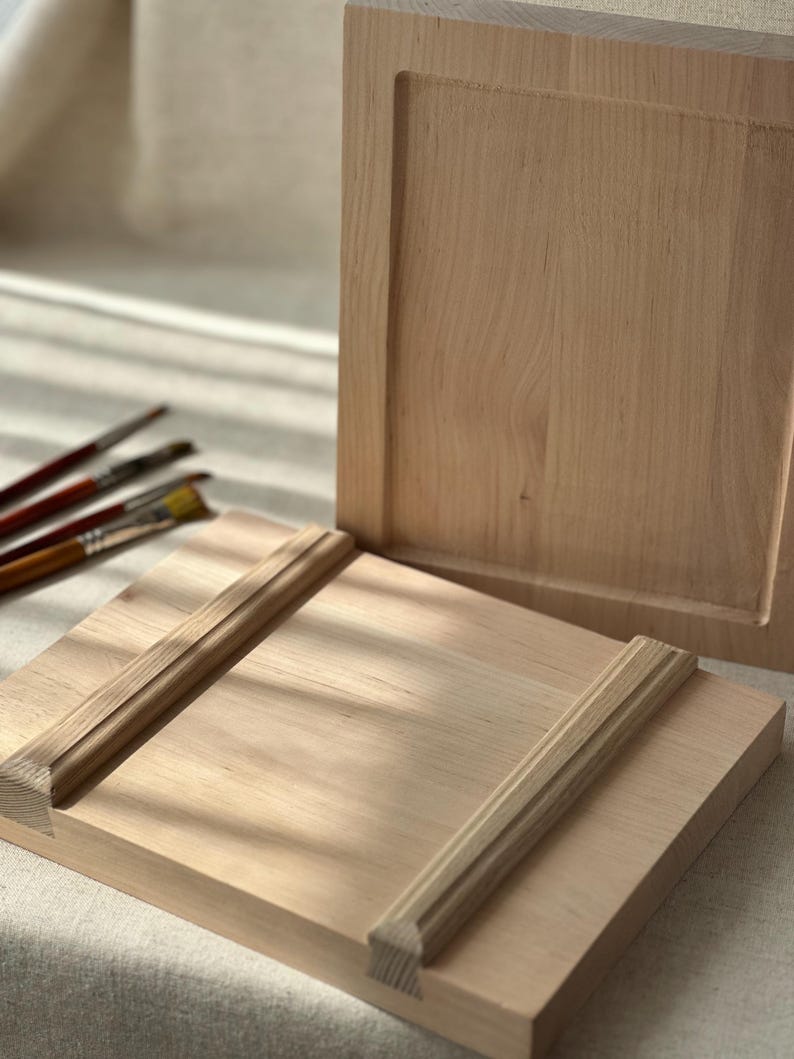 May include: Two unfinished wooden panels in a light brown hue. One panel features a recessed square, and the other has decorative trim. Paintbrushes are visible in the background, suggesting an art or craft theme.