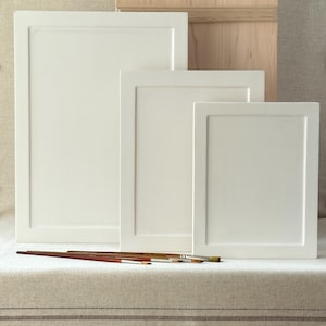 May include: A collection of white artist's canvases in varying sizes, arranged in front of a wooden box. Several paintbrushes are placed in front of the canvases. The items are set on a neutral-toned surface.