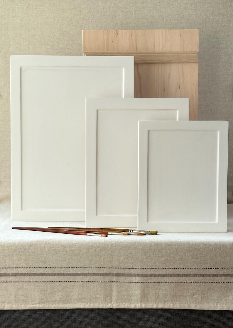 May include: A collection of white artist's canvases in varying sizes, arranged in front of a wooden box. Several paintbrushes are placed in front of the canvases. The items are set on a neutral-toned surface.