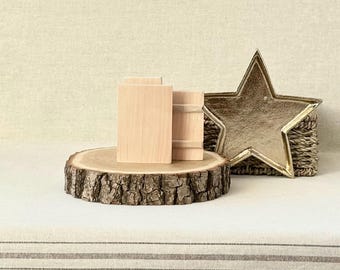 May include: A wooden tree slice with a natural bark edge supports three small, unfinished wooden blocks. A gold star-shaped decoration rests on a woven basket. The scene is set on a striped fabric.