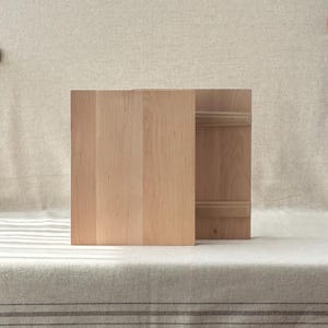 May include: A light-coloured wooden box with a lid, possibly for storage or display. The box is made of vertical wooden planks and sits on a neutral-toned surface. The lid has horizontal wooden slats.