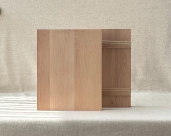 May include: A light-colored wooden box with a lid, likely for storage or display. The box is made of vertical wooden planks and sits on a neutral-toned surface. The lid has horizontal wooden slats.