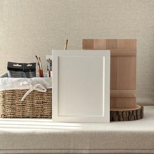 May include: A still-life composition featuring art supplies. A white framed panel stands next to a wooden block on a wood slice. A wicker basket with art supplies and paintbrushes is in the background. The scene is set on a striped linen surface.