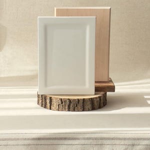 Può includere: Un piatto rettangolare in ceramica bianco sporco è esposto su una base di legno rustico, con un blocco di legno chiaro dietro. Il piatto è centrato su una sezione trasversale di un tronco d'albero, creando un'estetica naturale e minimalista.