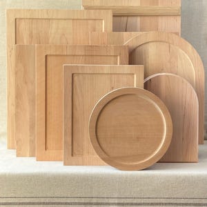 May include: A selection of wooden chopping boards and serving platters in various shapes. The items are crafted from light-coloured wood and include rectangular, square, and circular designs. Some have raised edges or decorative features.