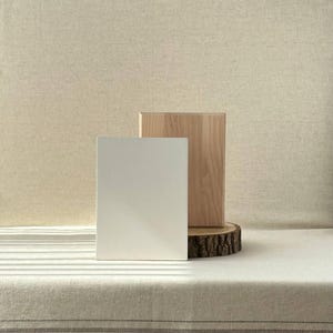 Könnte beinhalten: Minimalistische Produktpräsentation mit einem weißen rechteckigen Block und einem Naturholzblock, beide aufrecht stehend. Der Holzblock steht auf einer Baumscheibe. Der Hintergrund ist ein neutraler beiger Stoff, der eine saubere, moderne Ästhetik erzeugt.
