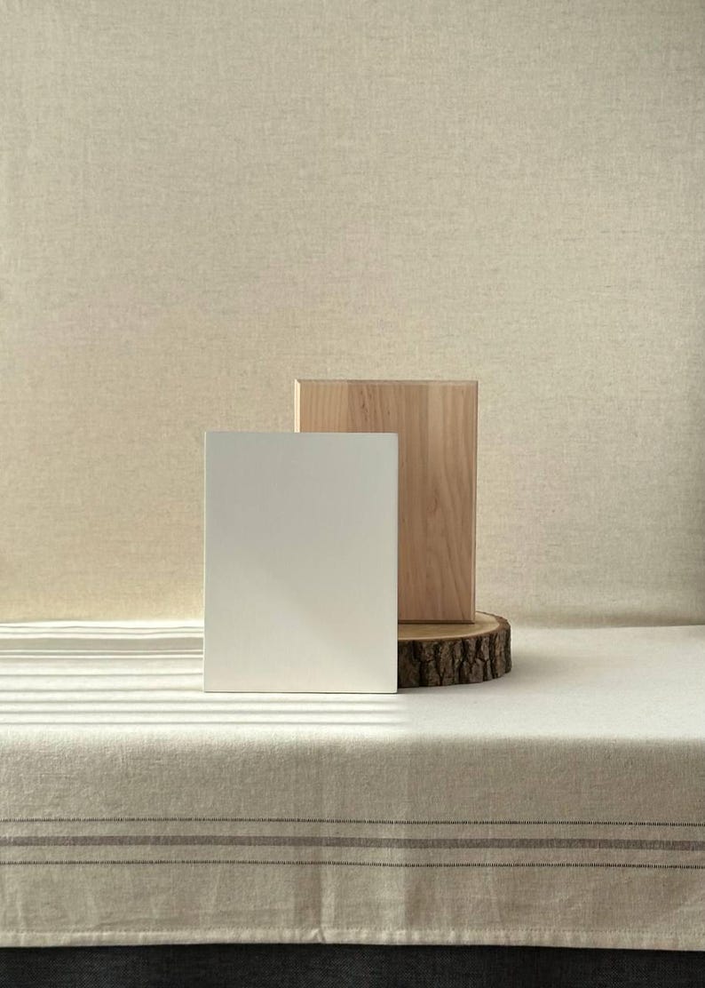 May include: A minimalist product display featuring a white rectangular block and a natural wood block, both standing upright. The wood block rests on a tree slice. The backdrop is a neutral beige fabric, creating a clean, modern aesthetic.