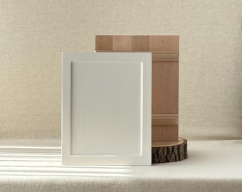 May include: A white, square cabinet door with a recessed panel stands next to a wooden panel and a wood slice. The cabinet door is painted a solid, matte white. The wooden panel has a natural wood grain.