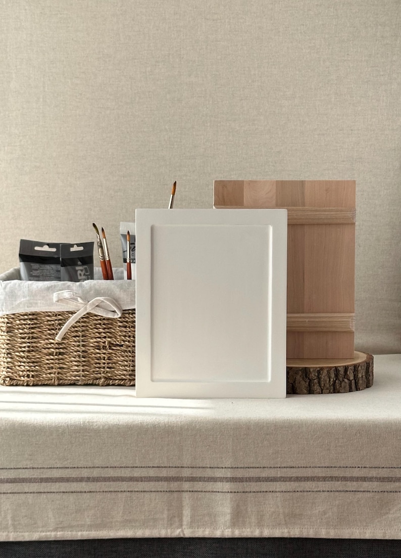 Pu&ograve; includere: Una natura morta con materiali per l'arte. Un pannello bianco incorniciato &egrave; posto accanto a un blocco di legno su una fetta di albero. Un cestino di vimini con fodera in lino contiene tubetti di vernice e pennelli. Lo sfondo &egrave; beige.