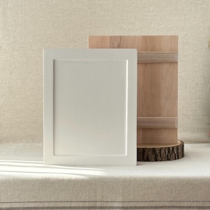 Può includere: Una porta bianca e rettangolare dell'armadio con un pannello incassato è accanto a un pannello di legno con cinghie orizzontali in legno. Il pannello di legno poggia su una piccola base rotonda di legno. Gli oggetti sono su una superficie di colore neutro.