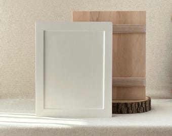 May include: A white, rectangular cabinet door with a recessed panel stands next to a wooden panel with horizontal wooden straps. The wooden panel rests on a small, round, wooden base. The objects are on a neutral-colored surface.