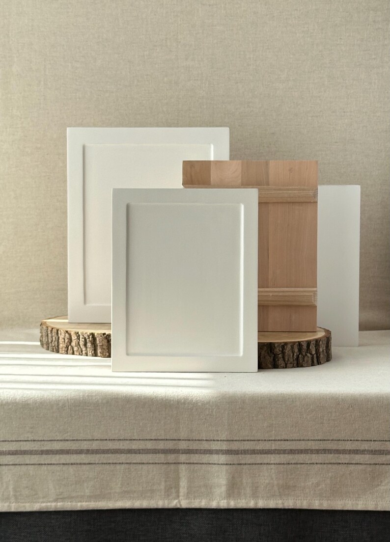 May include: An arrangement of cabinet door samples in various styles and finishes. Two white, framed doors stand prominently, alongside a natural wood door and a thin, white panel. The doors rest on a wooden slice, showcasing a rustic aesthetic.