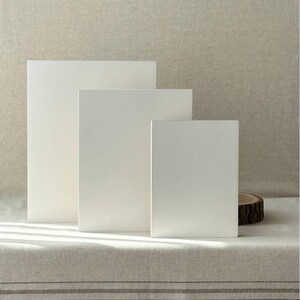 May include: Three white rectangular display blocks of varying sizes are arranged on a neutral-toned linen surface. The blocks stand upright, creating a minimalist and clean aesthetic. A small wooden slice is visible on the right.