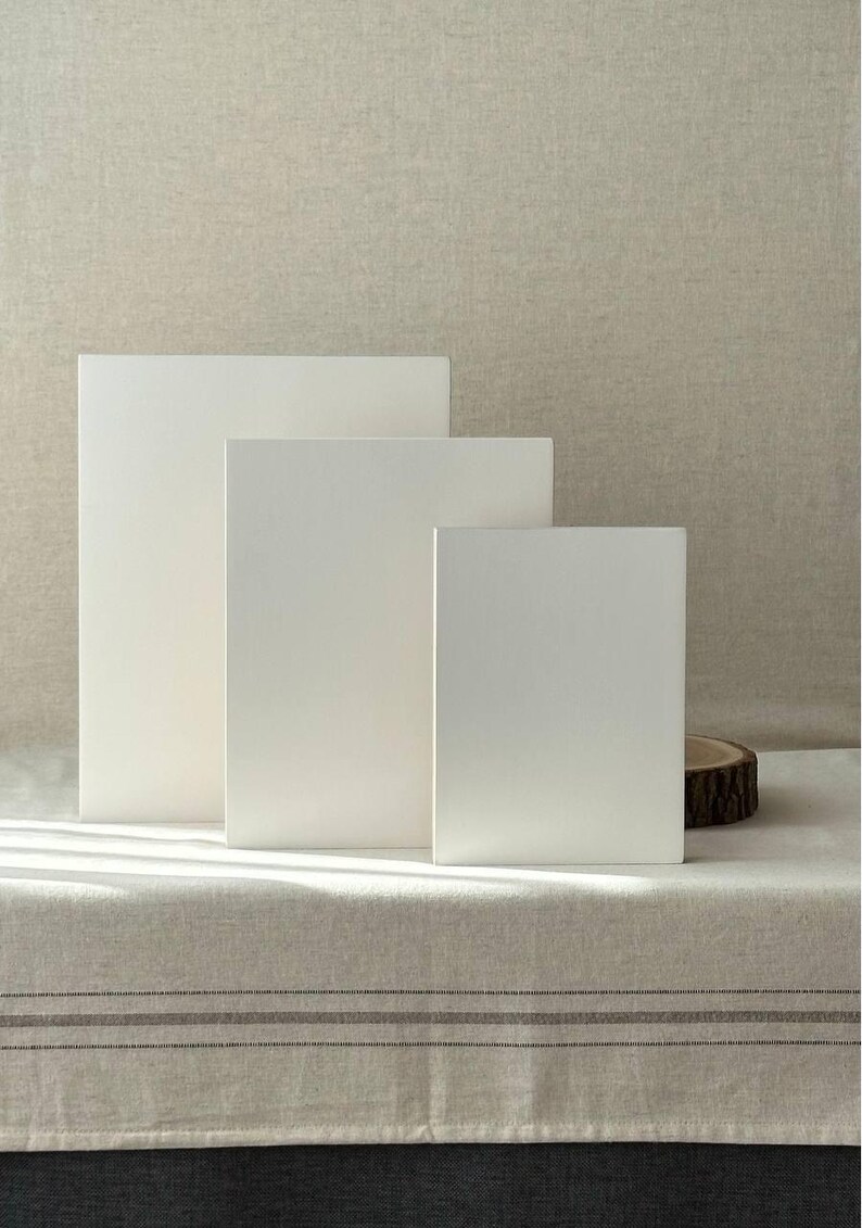 May include: Three white rectangular display blocks of varying sizes are arranged on a neutral-toned linen surface. The blocks stand upright, creating a minimalist and clean aesthetic. A small wooden slice is visible on the right.