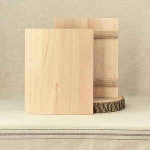 May include: Two light-coloured wooden cutting boards. One board stands upright, with a horizontal wooden handle. The other board is rectangular and lies flat in front. The boards are on a light-coloured surface.