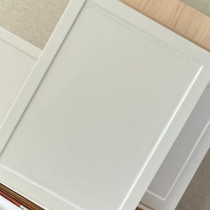 May include: Two white, rectangular art panels with a recessed border are stacked at a slight angle. The panels are resting on a piece of wood and a natural-colored fabric. Paint brushes are in the foreground.