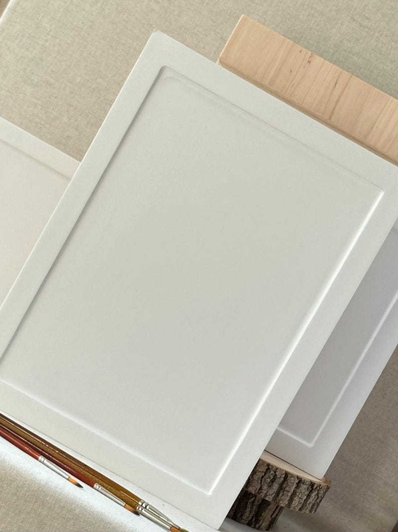 May include: Two white, rectangular art panels with a recessed border are stacked at a slight angle. The panels are resting on a piece of wood and a natural-colored fabric. Paint brushes are in the foreground.
